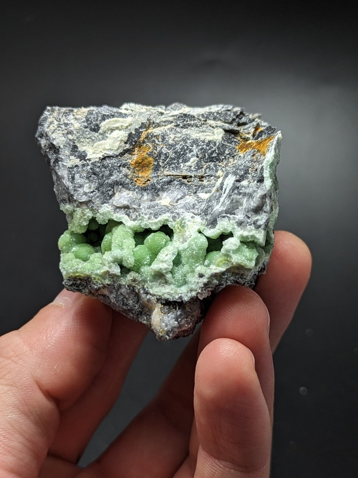A hand holds a striking green mineral specimen with a jagged, dark gray exterior and vibrant lime-green crystalline formations along its lower edge.