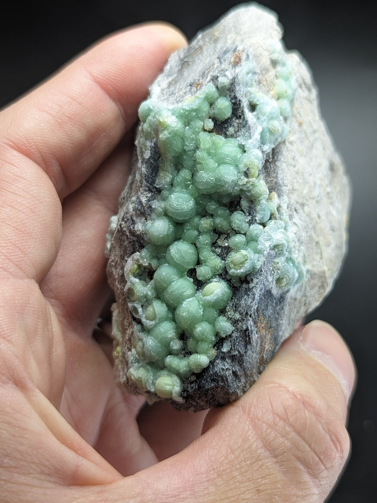 A hand holds a rough, grayish rock specimen featuring clusters of translucent, light green spherical crystals.