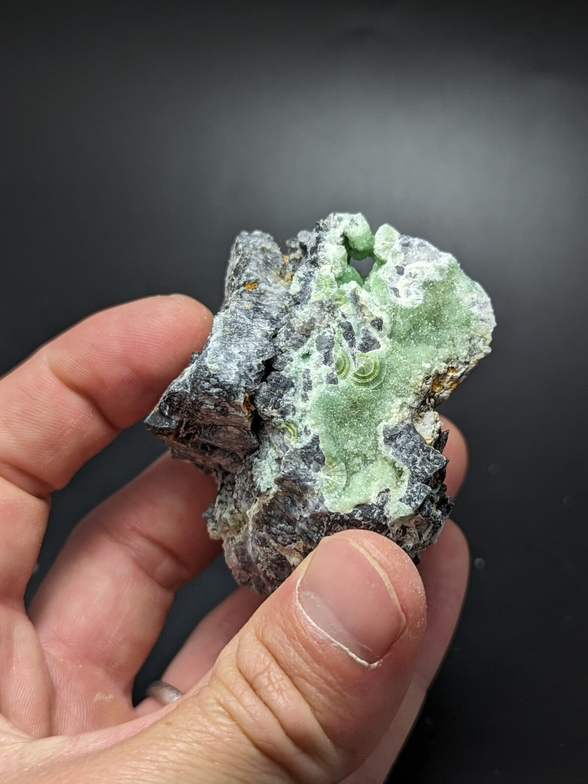A hand holds a rough, irregularly shaped mineral specimen featuring patches of vibrant green crystals embedded in a dark, textured matrix.
