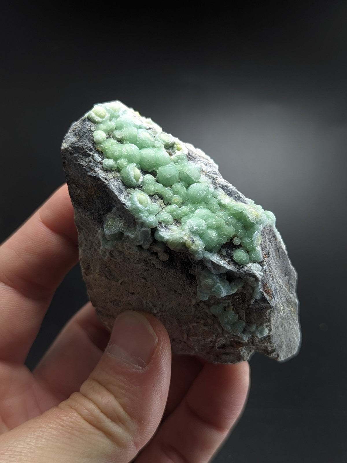 A hand holds a rough, gray rock specimen featuring clusters of vibrant light green, translucent crystals.
