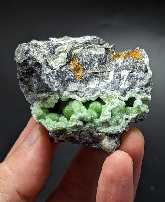 A hand holds a striking geological specimen featuring vibrant green, bead-like crystals nestled within a rugged matrix of gray and white rock, accented with patches of orange mineral.