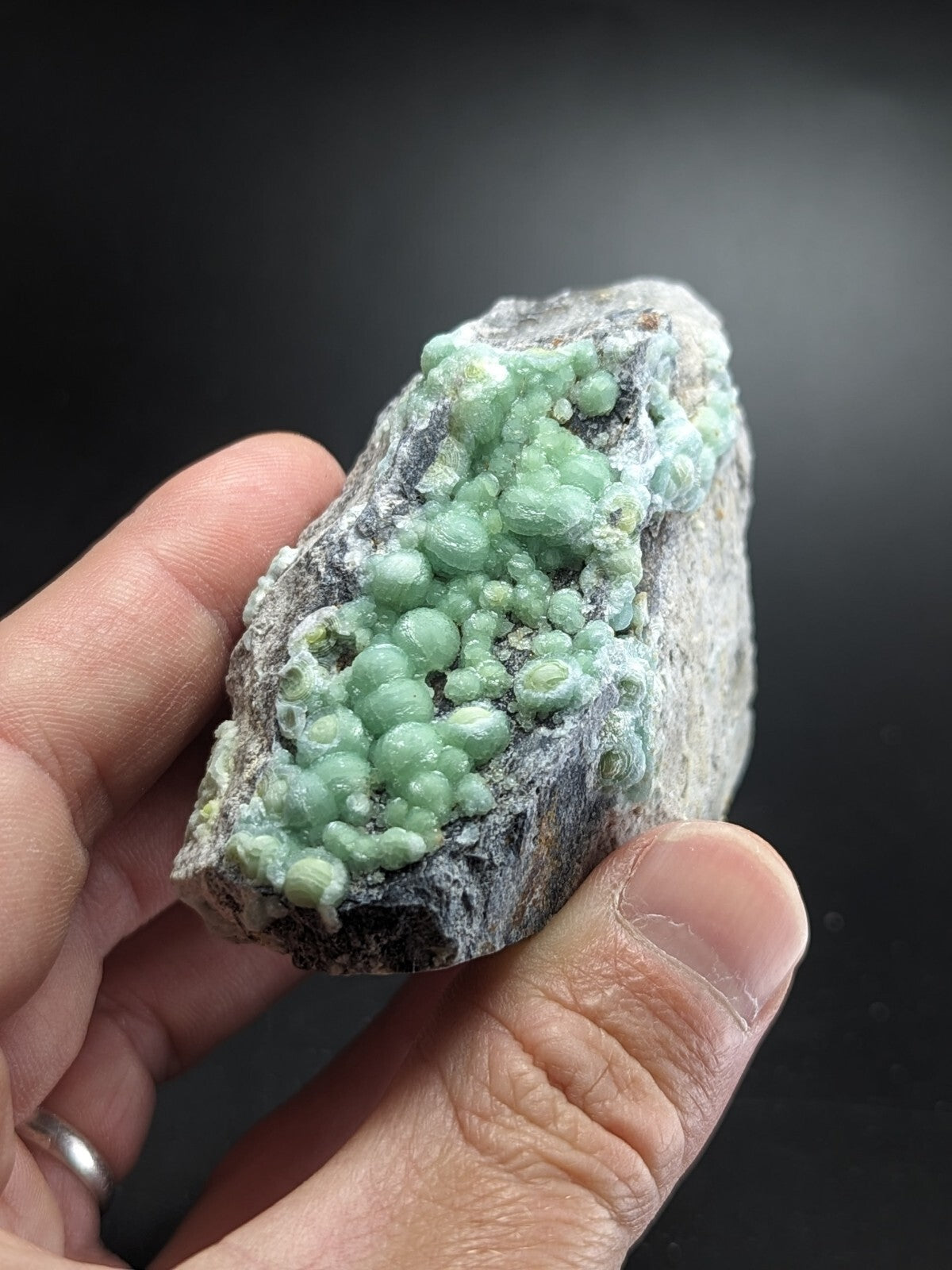 A hand holds a geological specimen featuring clusters of vibrant green, spherical crystals growing from a rough, grayish rock matrix.