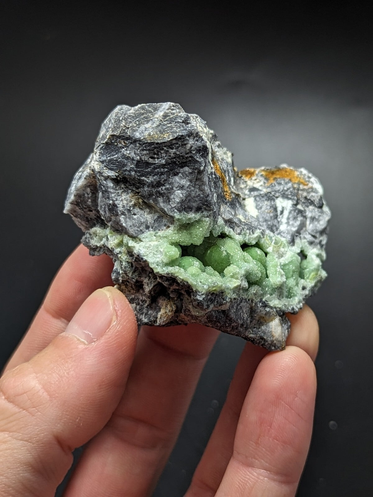 A hand holds a natural mineral specimen featuring vibrant green, rounded crystals emerging from a dark gray rock matrix with subtle orange streaks.