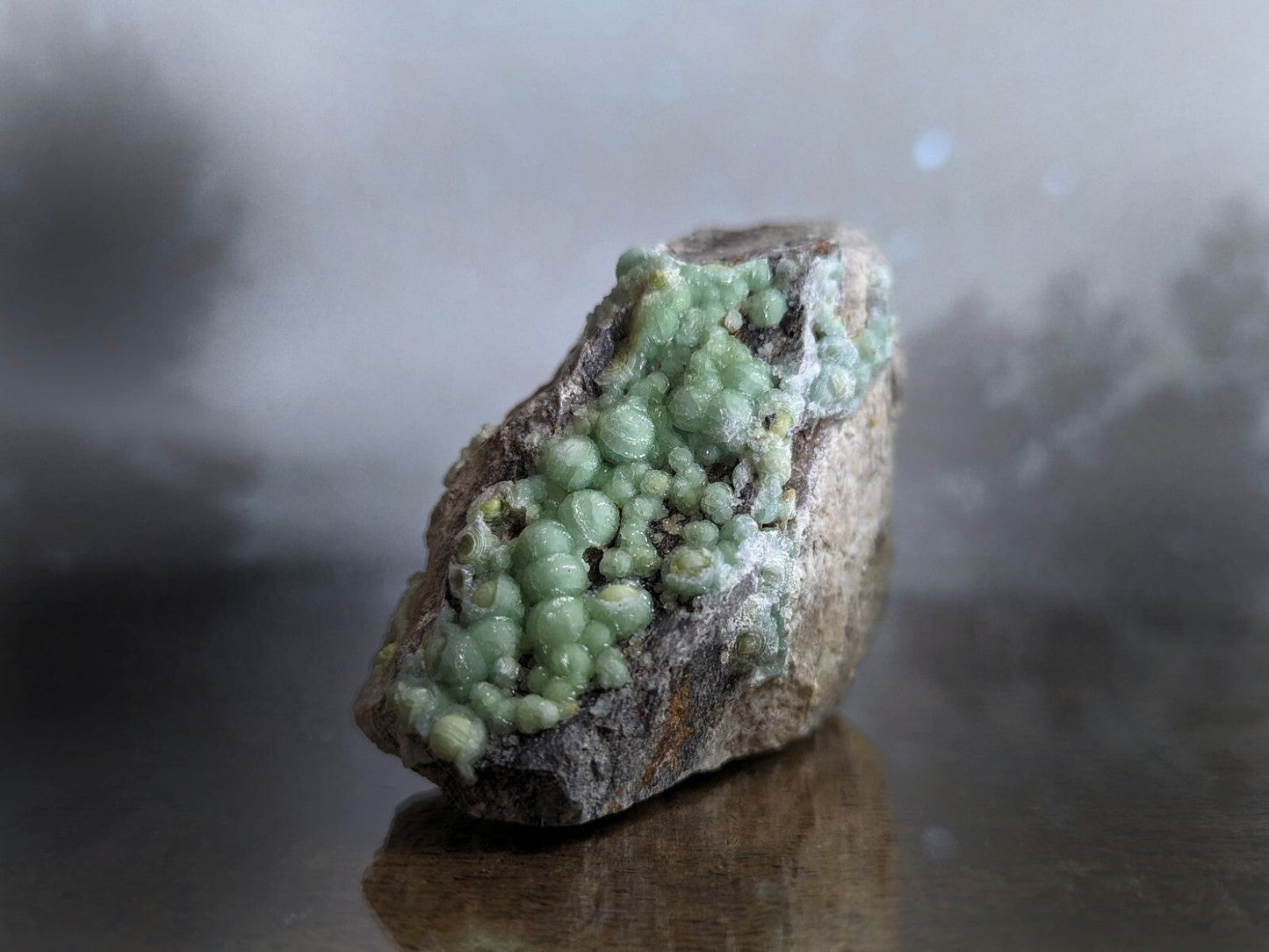 A rough, dark gray rock with a cluster of translucent green spherical crystals growing from its surface.