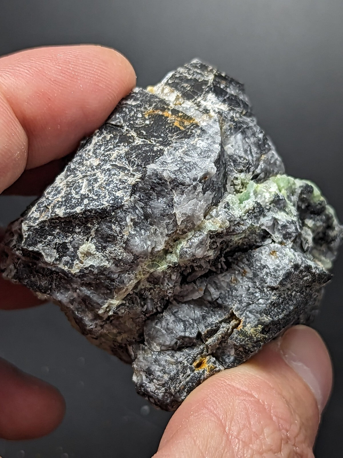 A rugged, irregularly shaped mineral specimen with a dark gray base and patches of greenish hues.