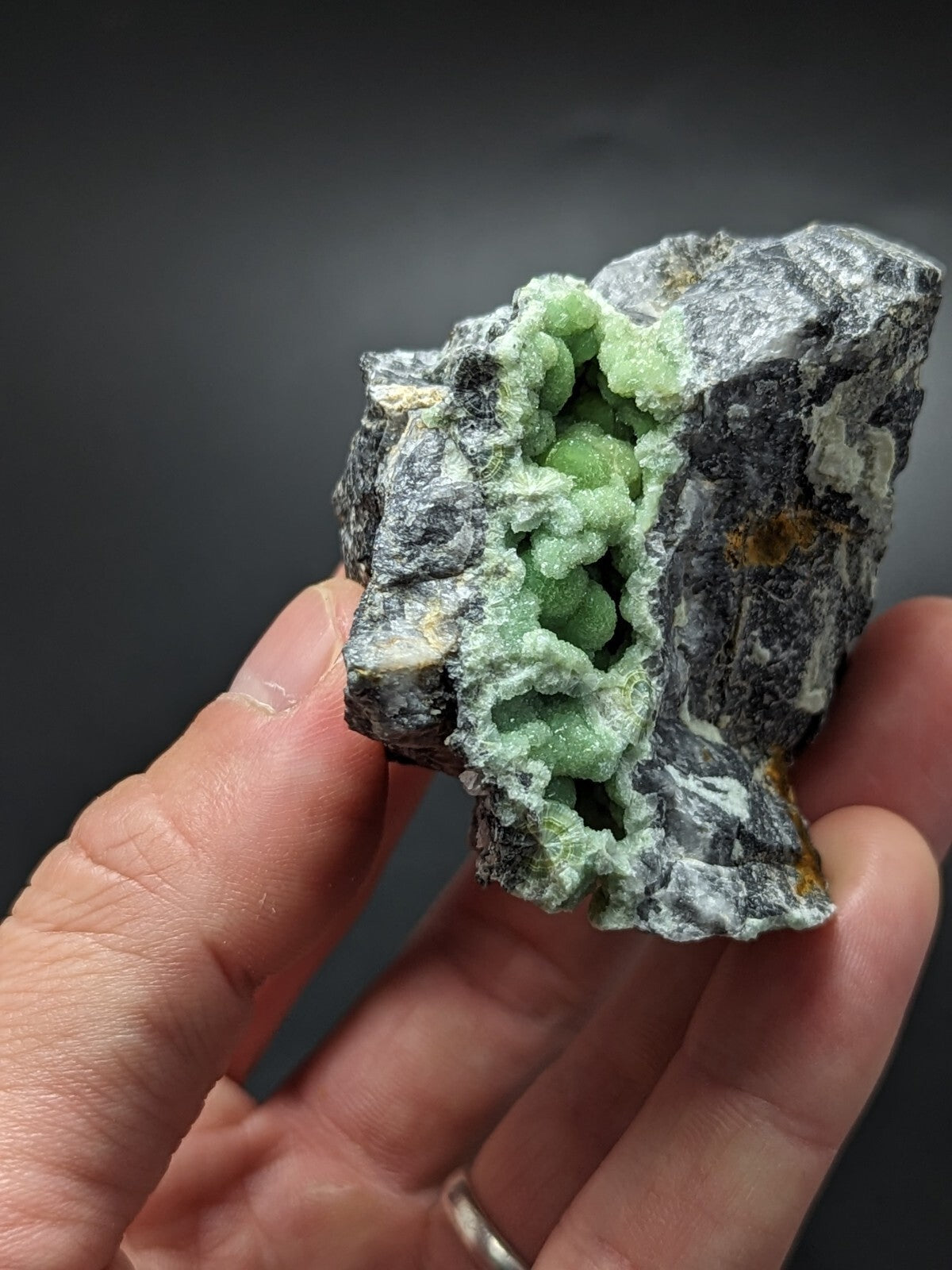 A hand holds a striking green druzy geode with its inner crystalline structure exposed against a dark, rocky exterior.