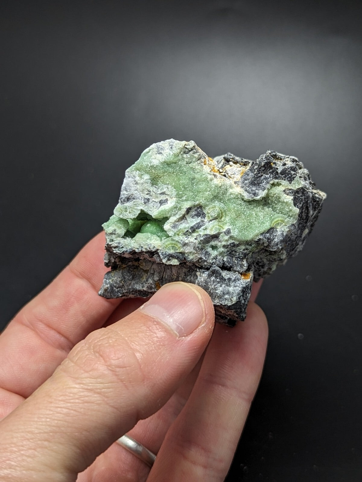 A hand holds a rough, irregularly shaped mineral specimen featuring vibrant green crystalline formations set against a dark gray matrix with subtle golden flecks.