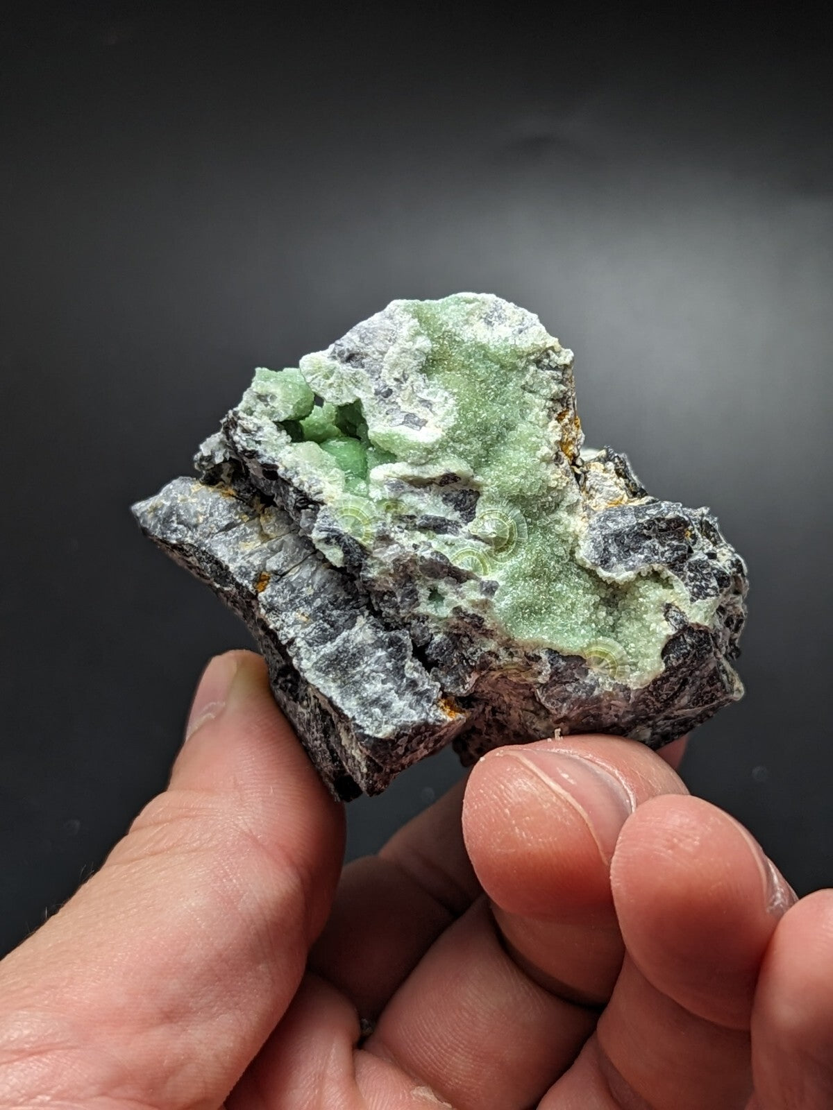 A hand holds a rough, irregularly shaped mineral specimen featuring vibrant green crystalline formations set against a dark gray matrix.