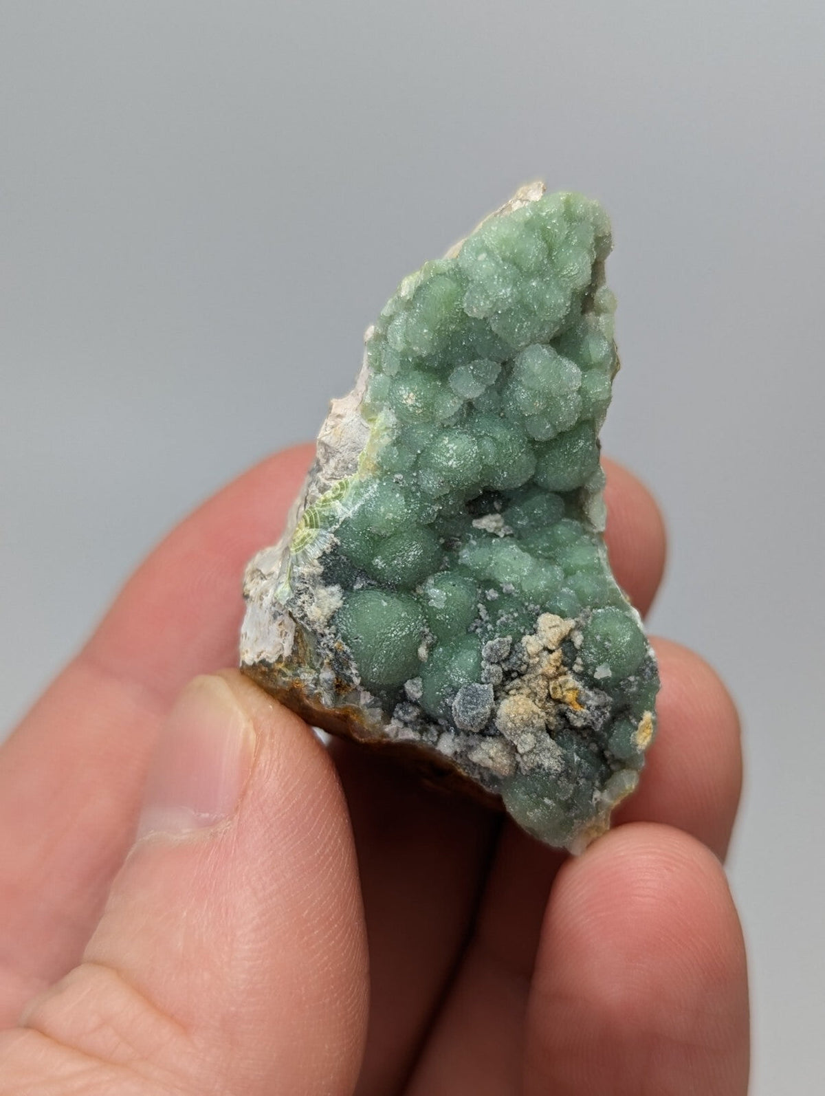 A hand holds a small, irregularly shaped mineral specimen featuring clusters of translucent green crystals growing from a rougher matrix with patches of white and yellowish material.