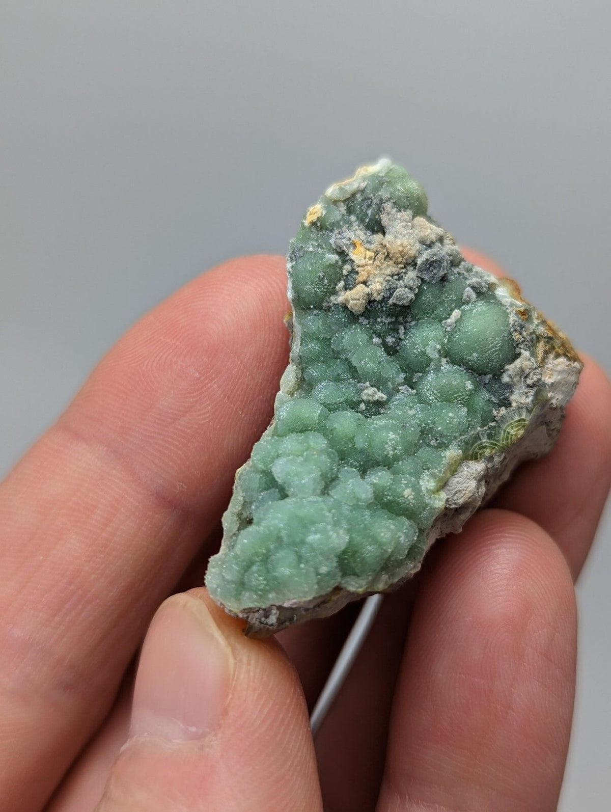 A hand holds a rough, light green mineral specimen with rounded nodules and patches of white and yellowish matrix.