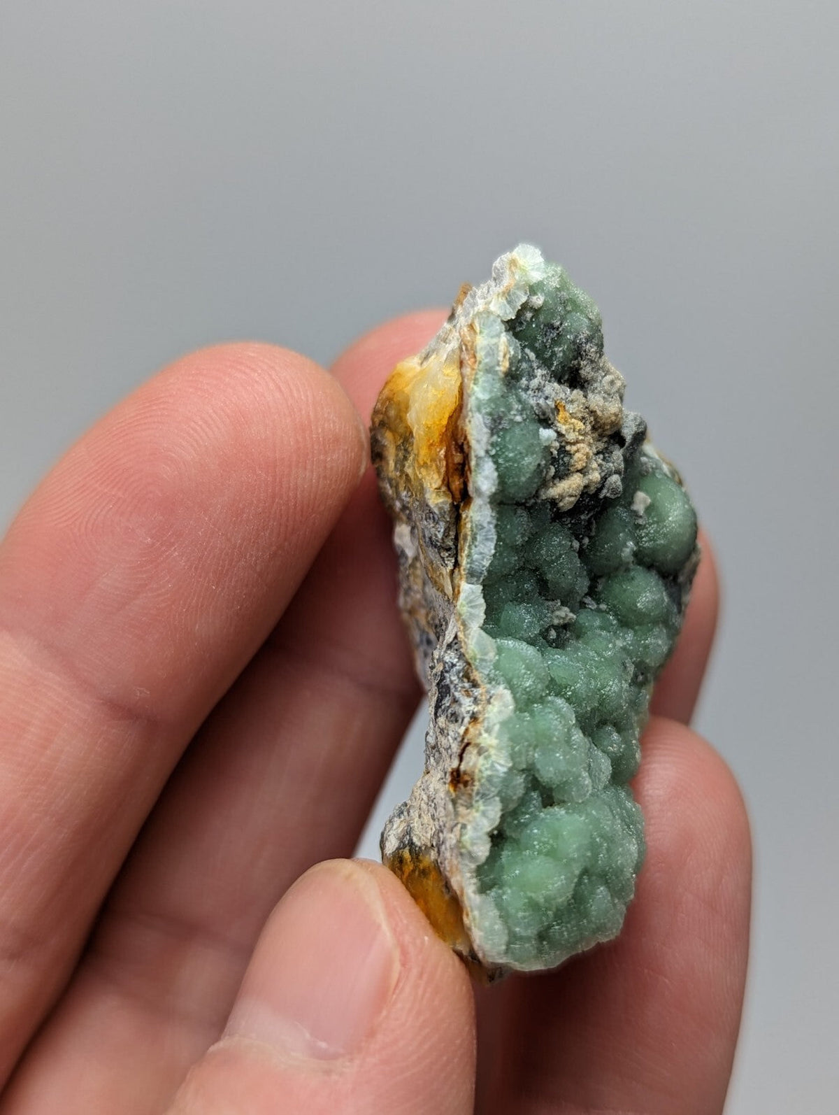 A hand holds a small, irregularly shaped mineral specimen featuring vibrant green crystalline formations with patches of yellow and white matrix material.