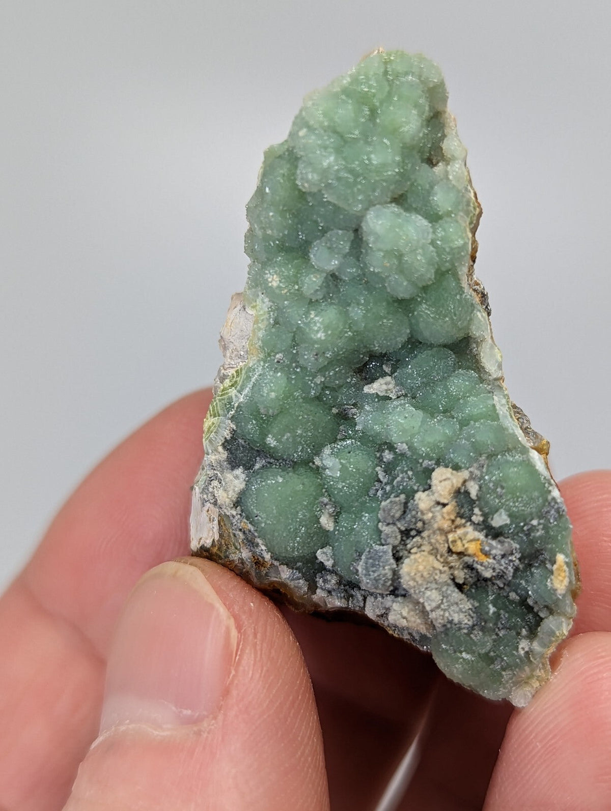 A hand holds a green mineral specimen with clustered, rounded crystals forming a textured surface.