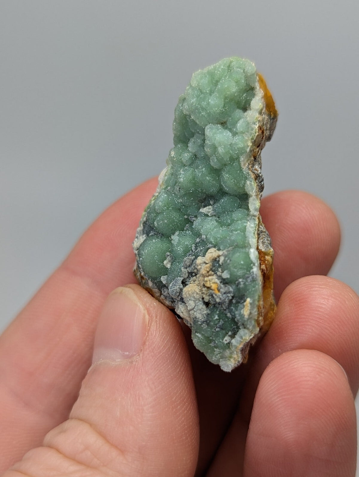 A hand holds a light green, bumpy mineral specimen with patches of white and yellowish material.