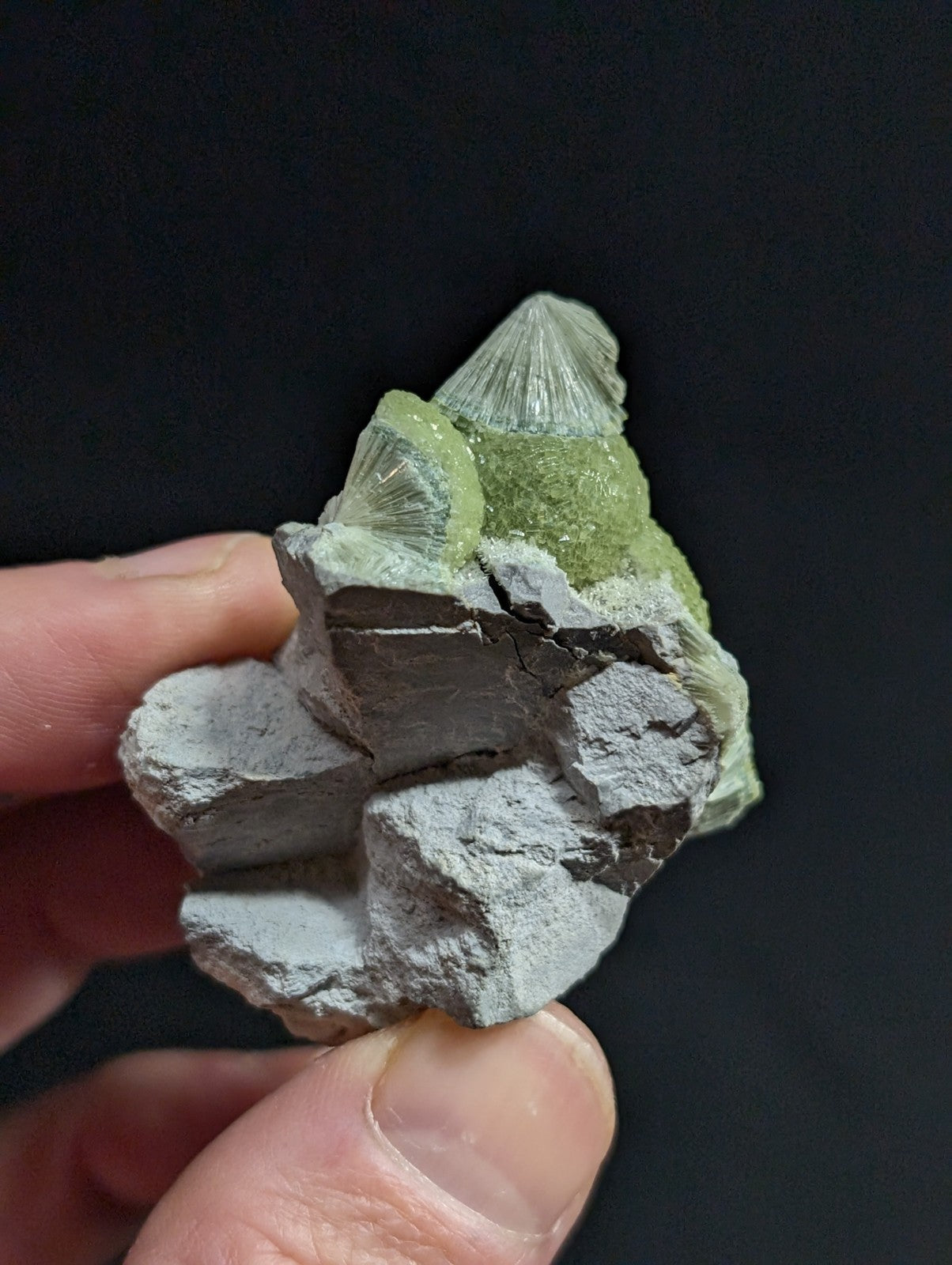 A hand holds a natural mineral specimen featuring translucent green crystals with radial facets growing from a rough, gray rock base.