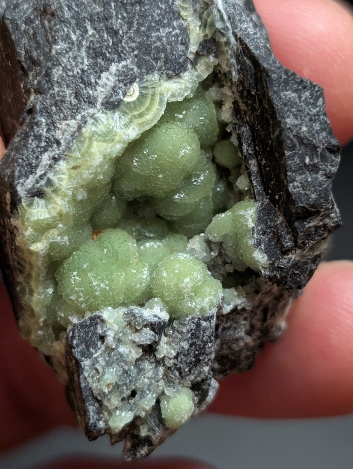 A hand holds a dark, rough-textured rock containing clusters of translucent green crystals.