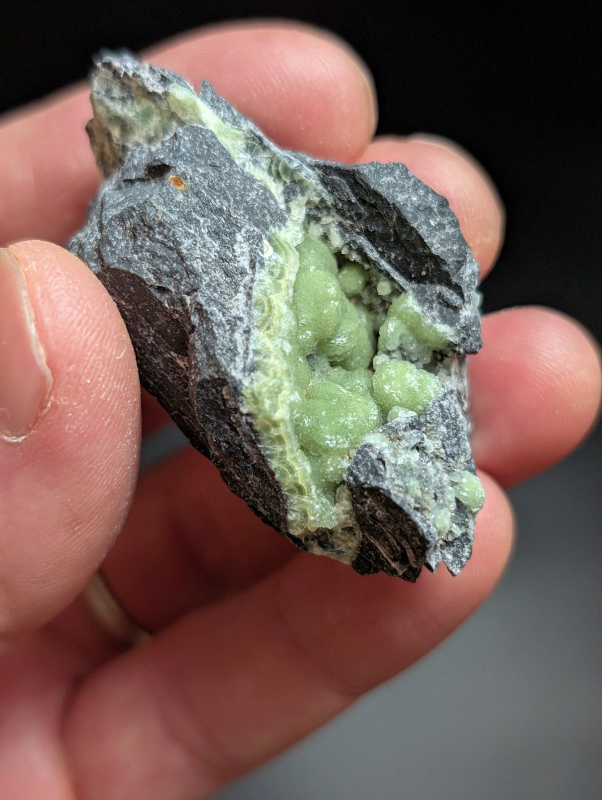 A hand holds a rough, dark gray rock with vibrant green mineral crystals embedded within its fractured surface.