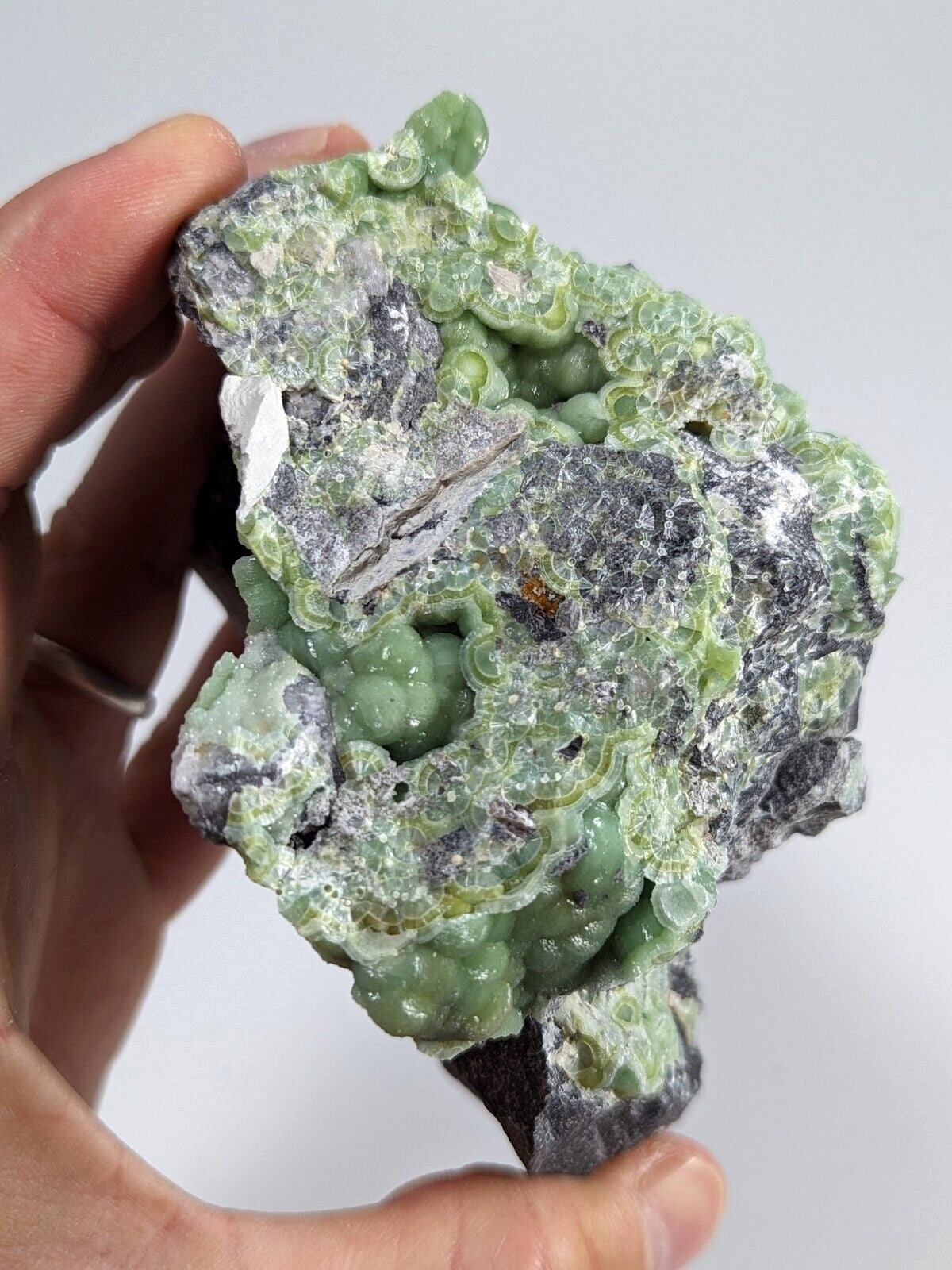 A hand holds a rough, irregularly shaped mineral specimen featuring vibrant green, bubble-like formations over a dark gray matrix.