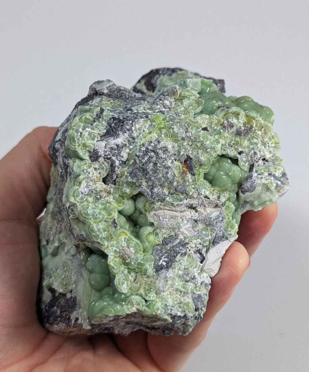 A hand holds a rough, irregularly shaped mineral specimen featuring clusters of translucent green spherical crystals embedded in a grayish matrix.