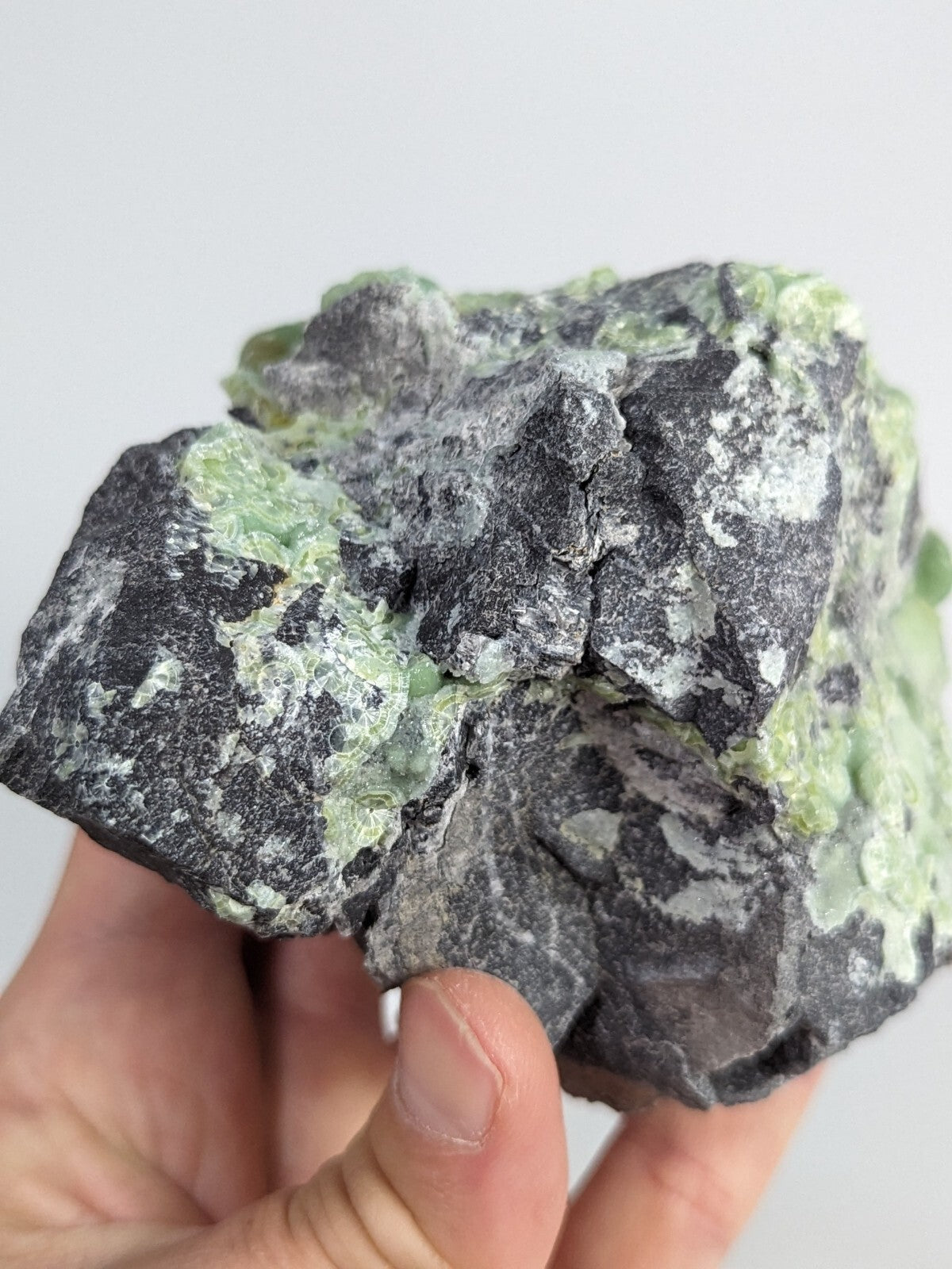 A rugged, dark gray rock specimen with patches of translucent green mineral crystals embedded across its uneven surface.