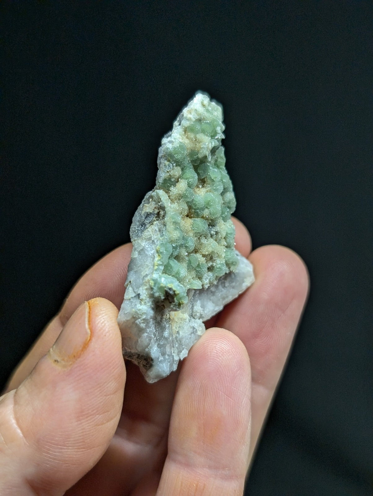 A hand holds a rough, jagged mineral specimen with translucent green crystals growing from a lighter gray base.