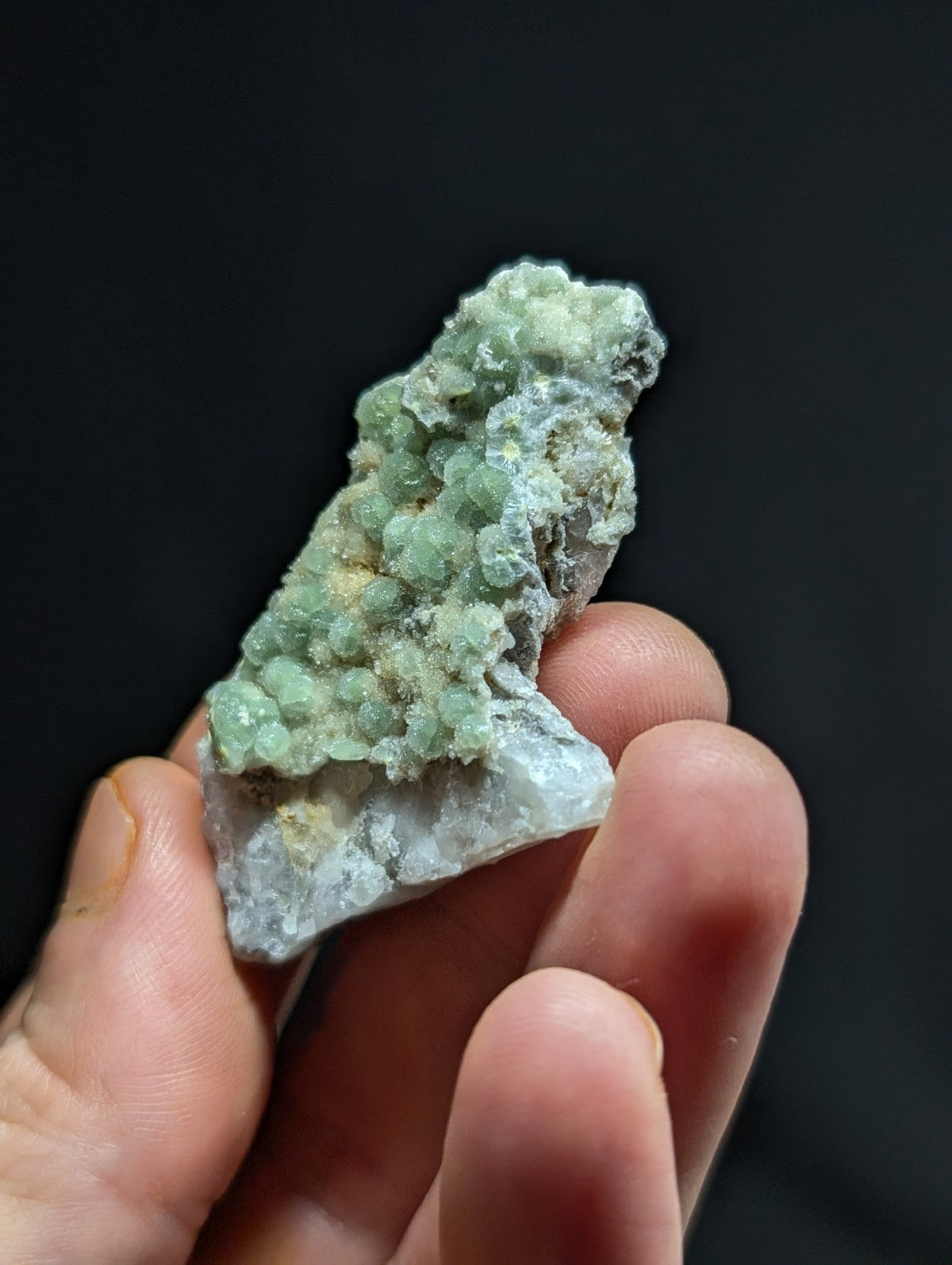 A hand holds a rough, irregularly shaped mineral specimen featuring clusters of translucent green crystals growing from a lighter grayish base.
