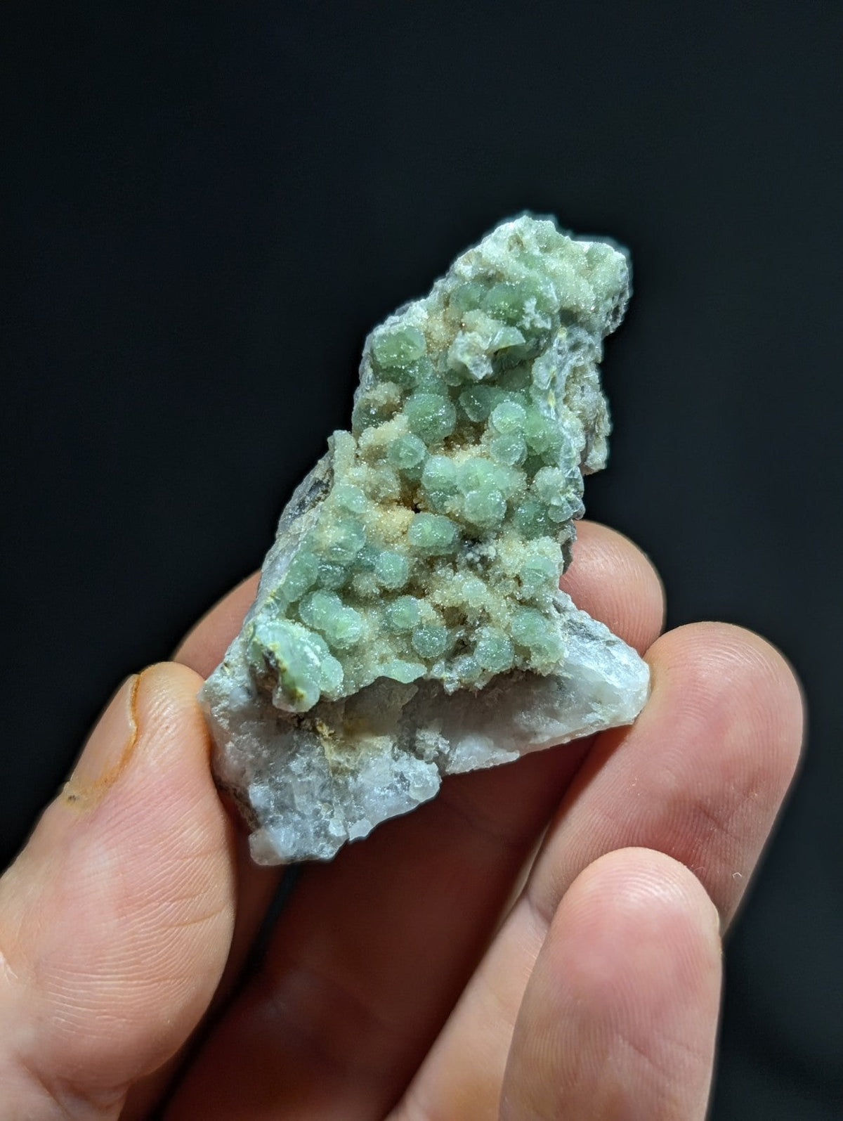 A hand holds a natural mineral specimen featuring clusters of translucent, light green crystals growing from a rougher, pale gray matrix.