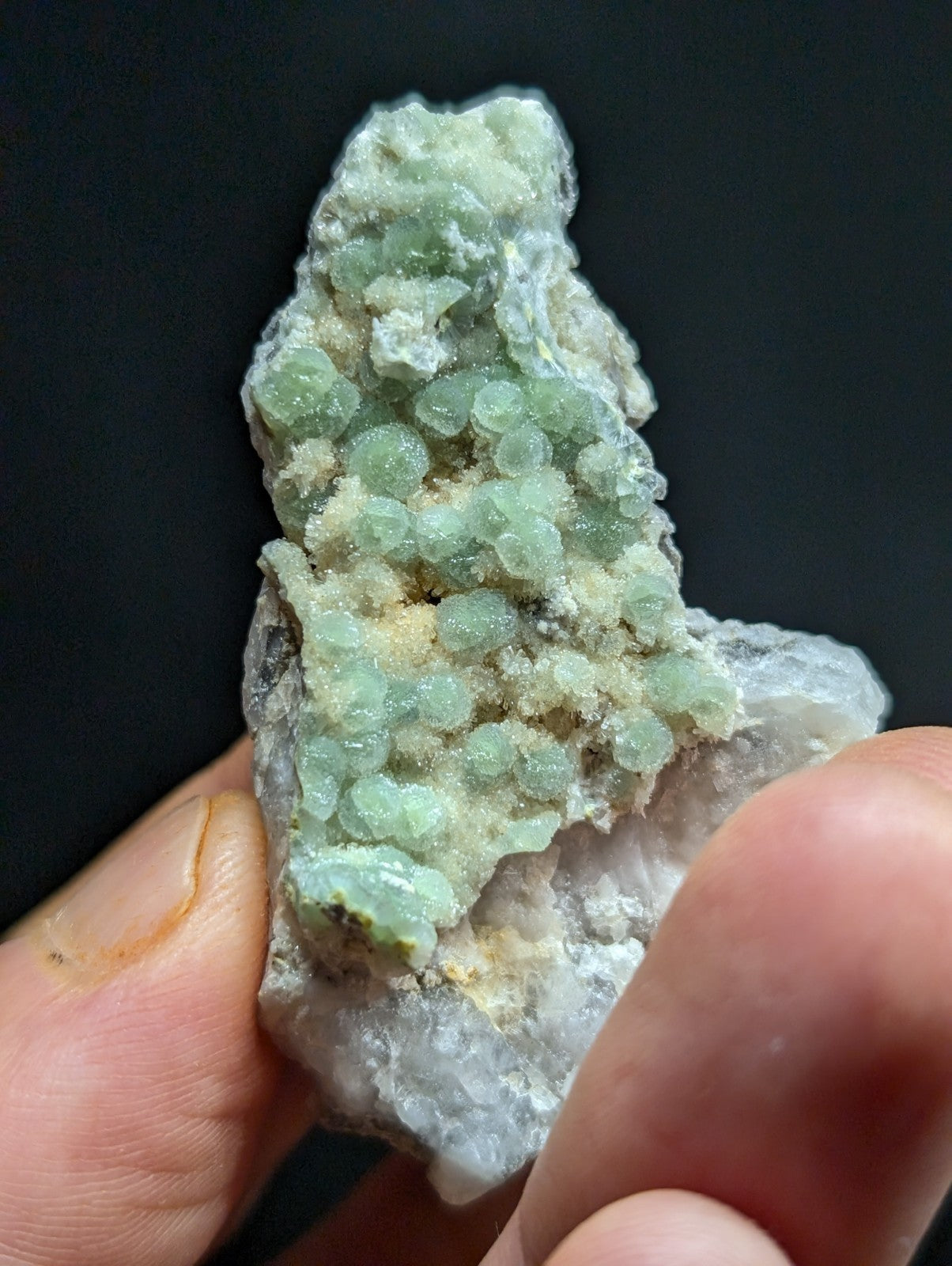 A hand holds a geological specimen featuring clusters of translucent, pale green spherical crystals growing on a white crystalline matrix.
