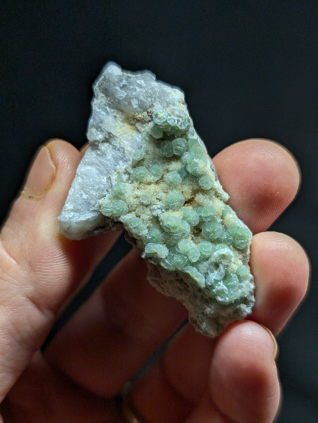 A hand holds a rough, irregularly shaped mineral specimen featuring clusters of translucent green crystals growing from a lighter gray matrix.