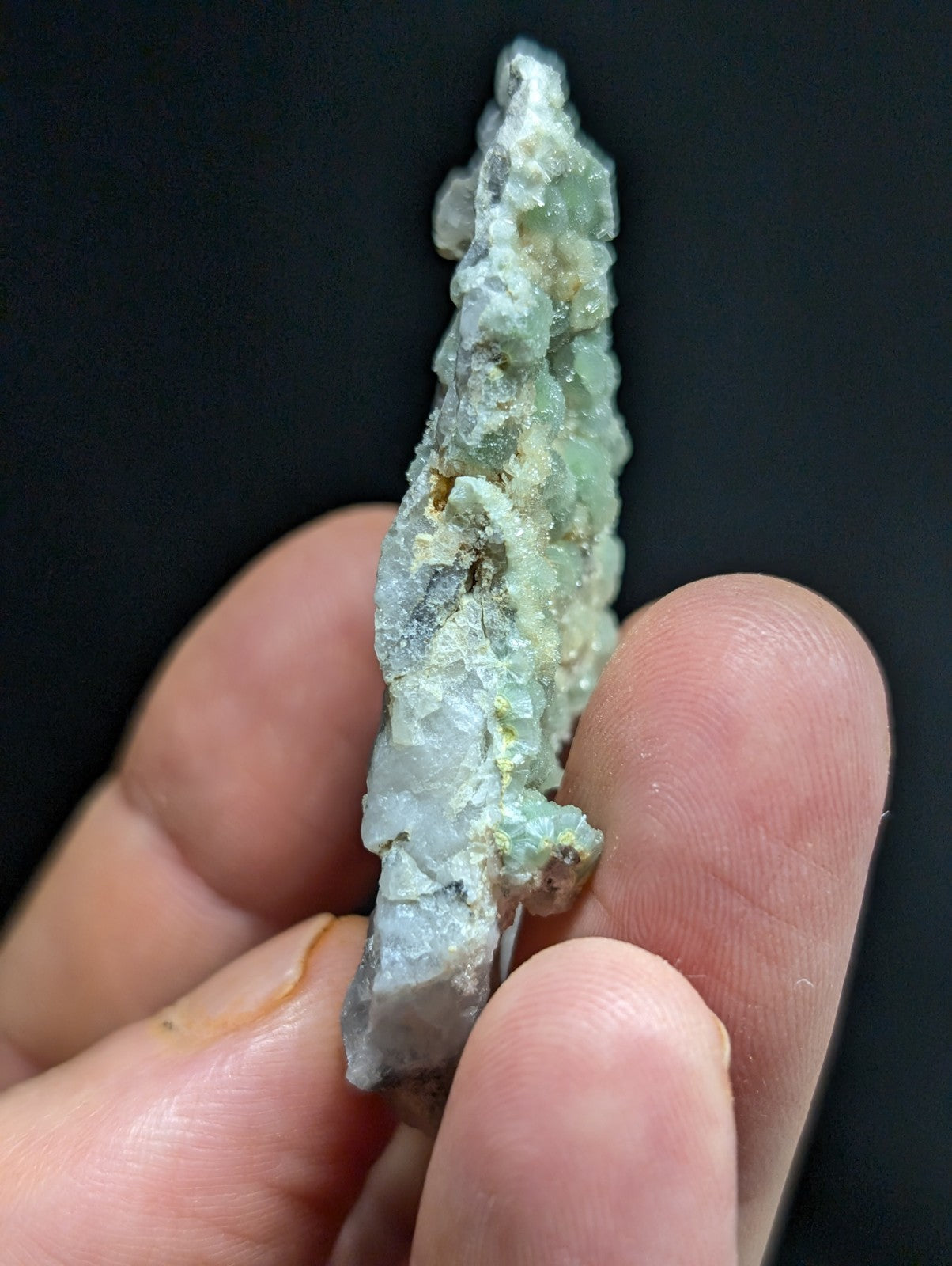 A translucent greenish-blue mineral specimen with crystalline textures and patches of yellowish material, held between fingers against a dark background.