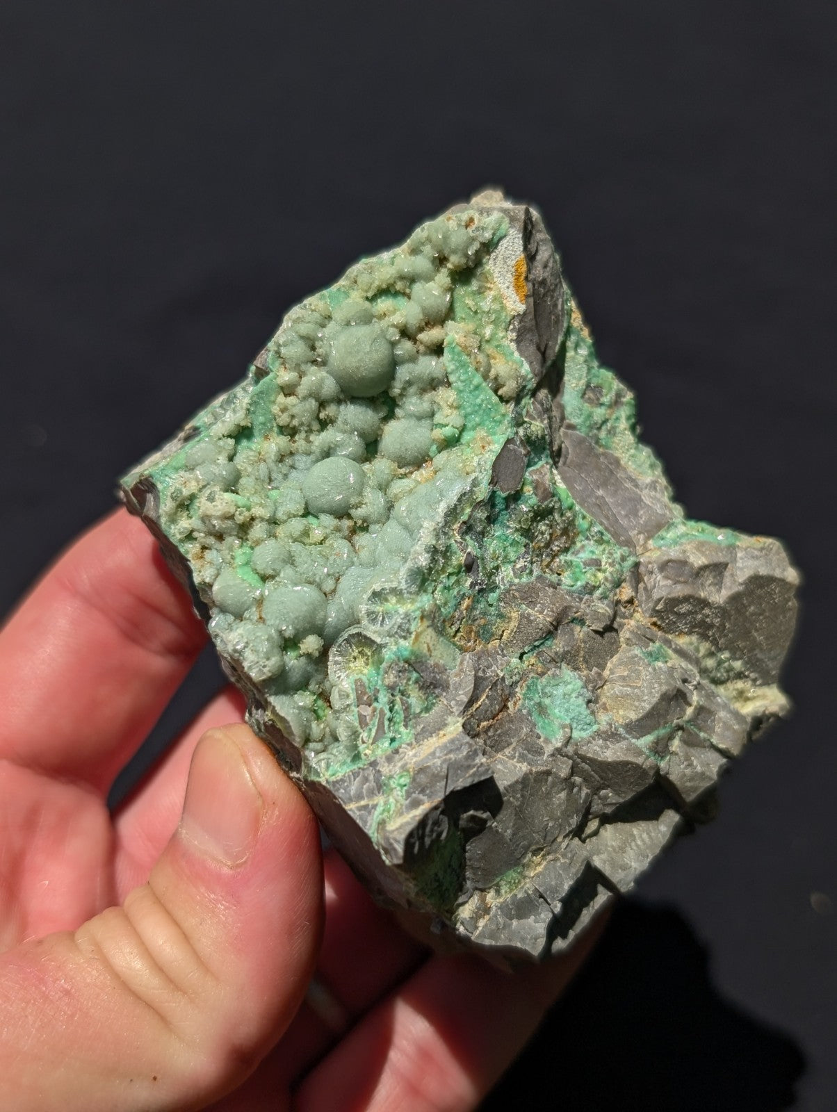 A hand holds a rough, irregularly shaped mineral specimen featuring vibrant green coppery crystals growing from a darker gray rock base.