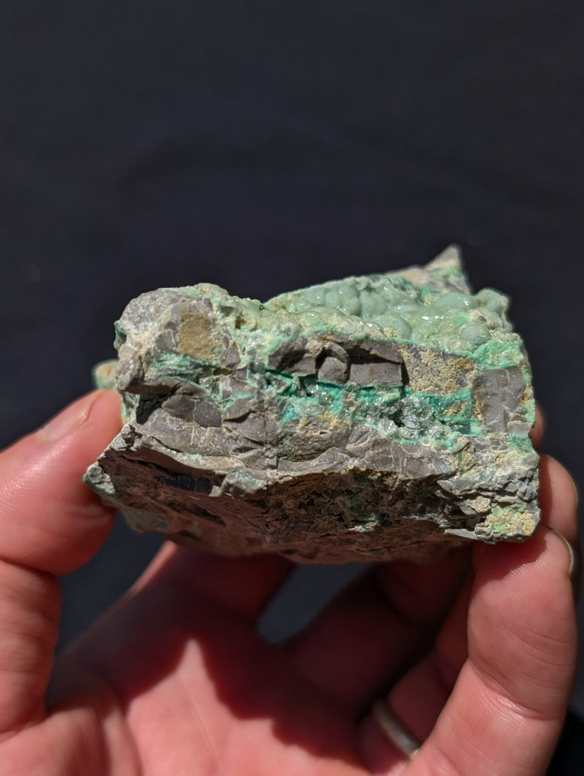 A rough, irregularly shaped piece of green malachite mineral with patches of darker gray rock and a visible vein running through its center.