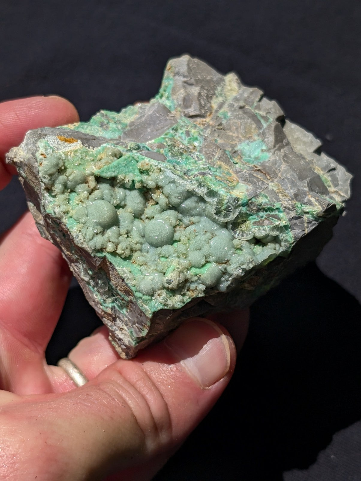 A hand holds a rough, irregularly shaped mineral specimen featuring vibrant green nodules and patches of grayish rock.