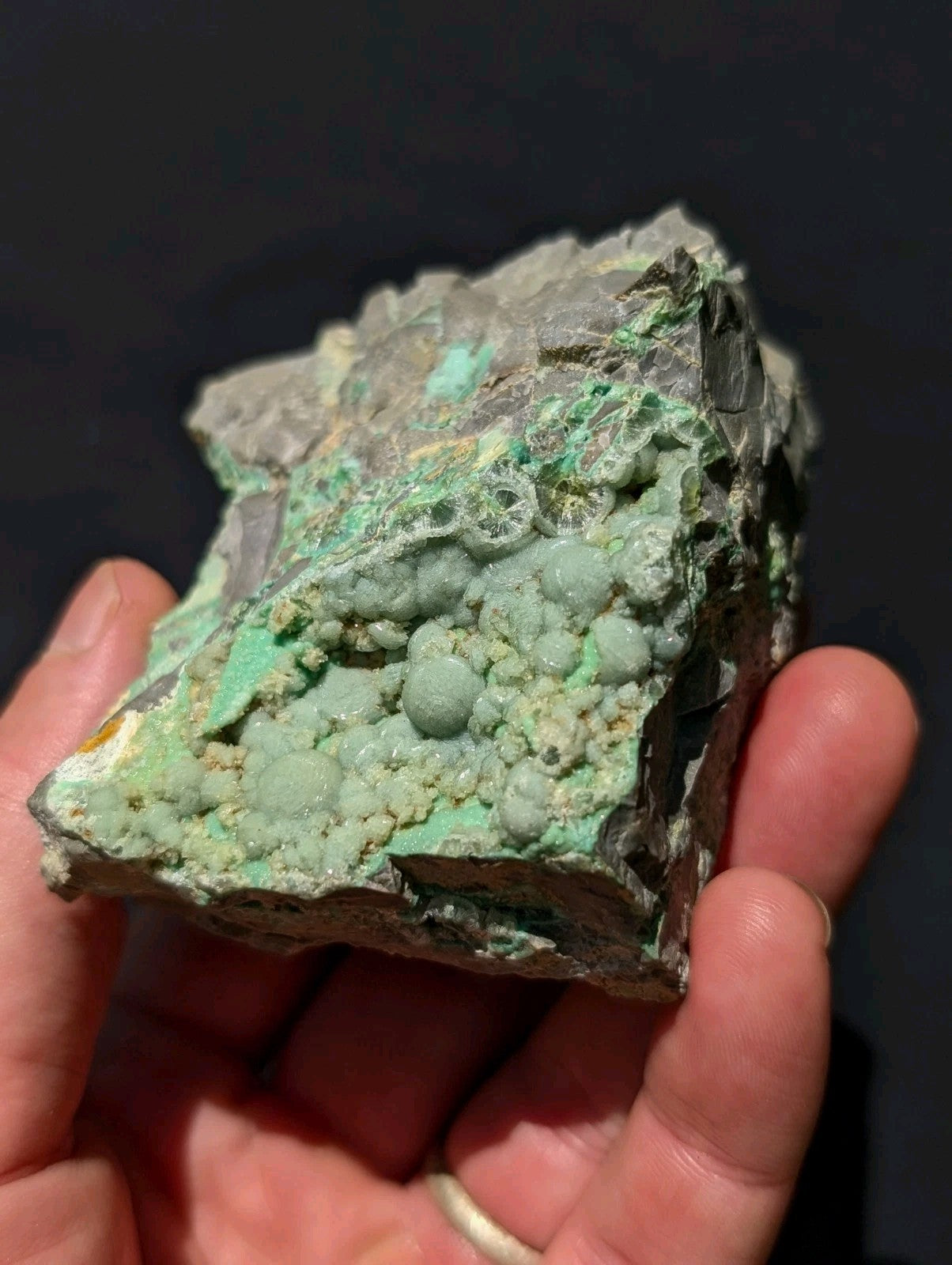 A hand holds a rough, irregularly shaped piece of greenish-blue malachite mineral with patches of gray matrix and small rounded nodules.