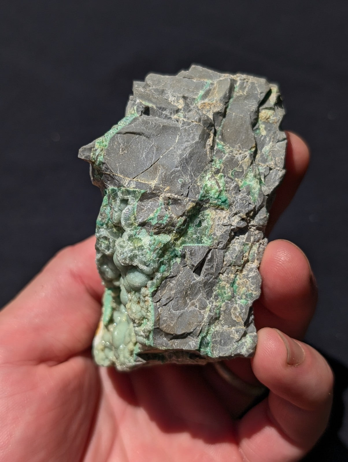 A hand holds a rough, irregularly shaped mineral specimen with a gray base and patches of vibrant green patina.