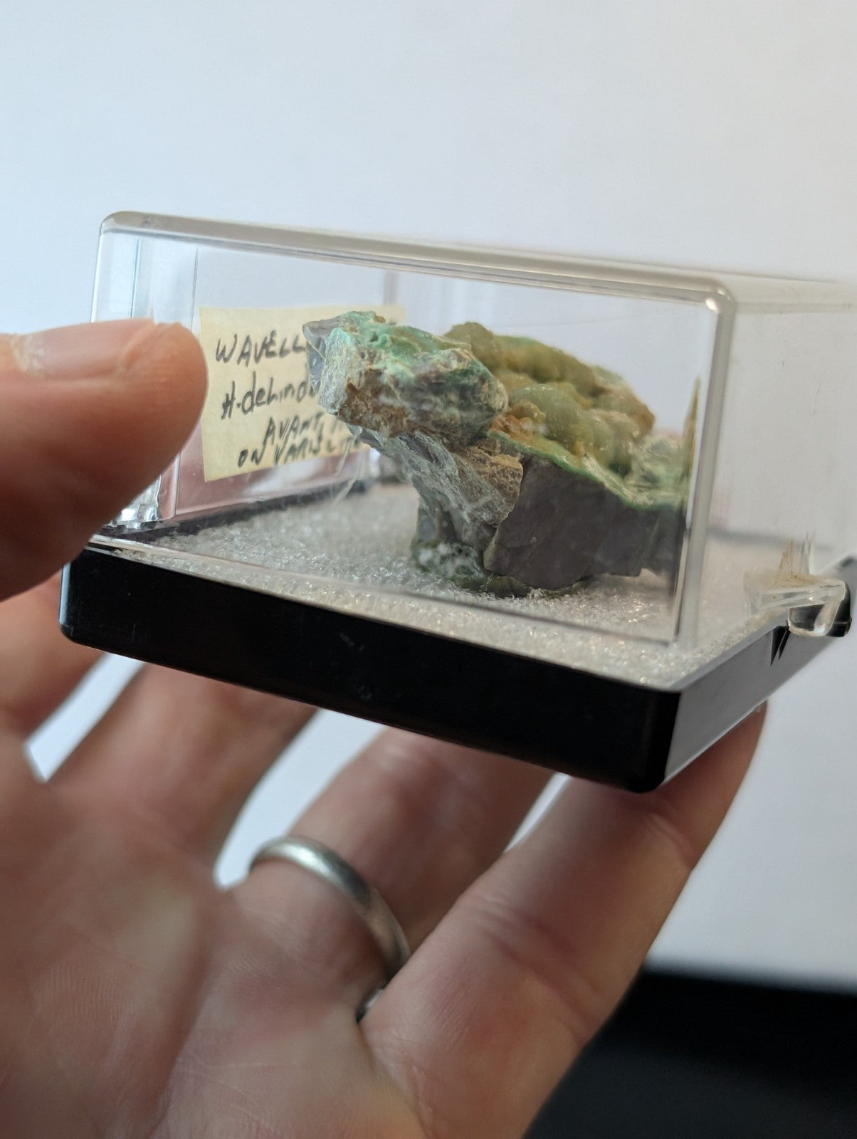 A green and brown mineral specimen displayed in a clear acrylic display case with a black base.