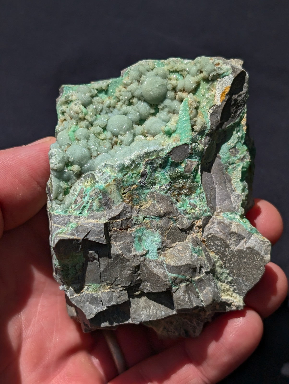 A hand holds a rough, irregularly shaped mineral specimen featuring vibrant green crystalline formations atop a darker gray rock base.