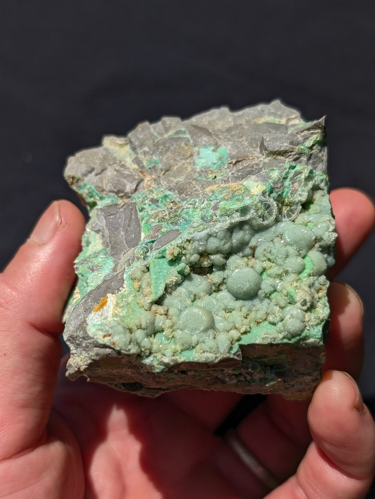 A hand holds a rough, irregularly shaped mineral specimen featuring vibrant green crystalline formations over a grayish matrix.