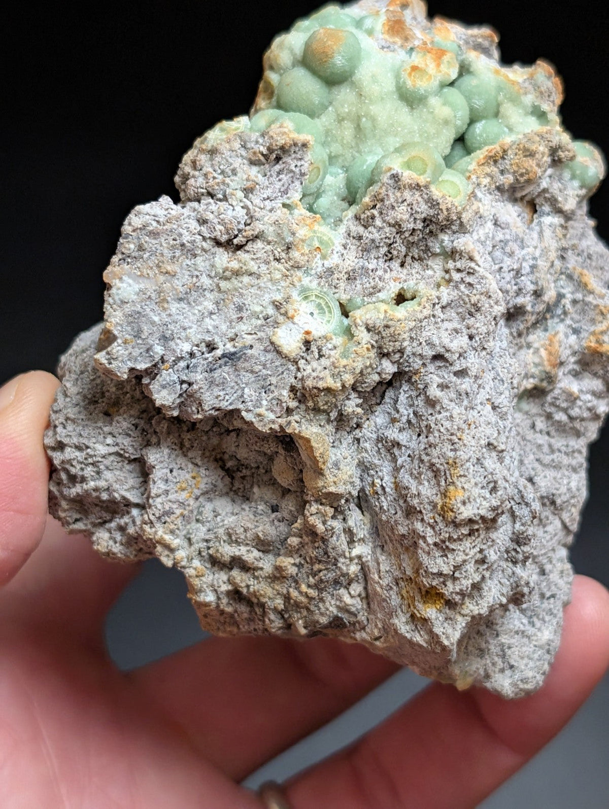 A hand holds a rough, grayish rock specimen featuring clusters of light green, bulbous crystals with hints of orange and yellow mineralization.