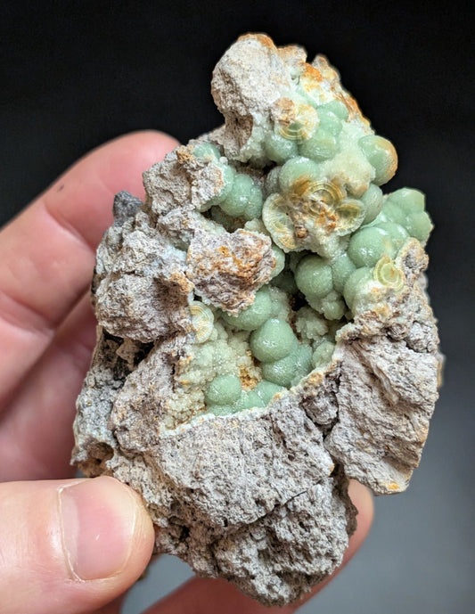 A hand holds a striking greenish-blue mineral specimen featuring rounded, bead-like formations nestled within a rough gray matrix.