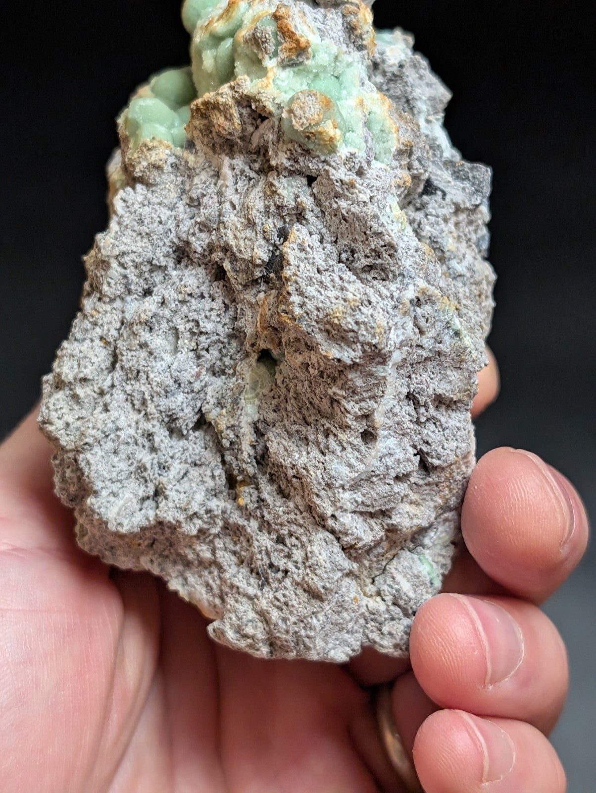 A hand holds a rough, grayish rock specimen featuring patches of vibrant green mineral growth.