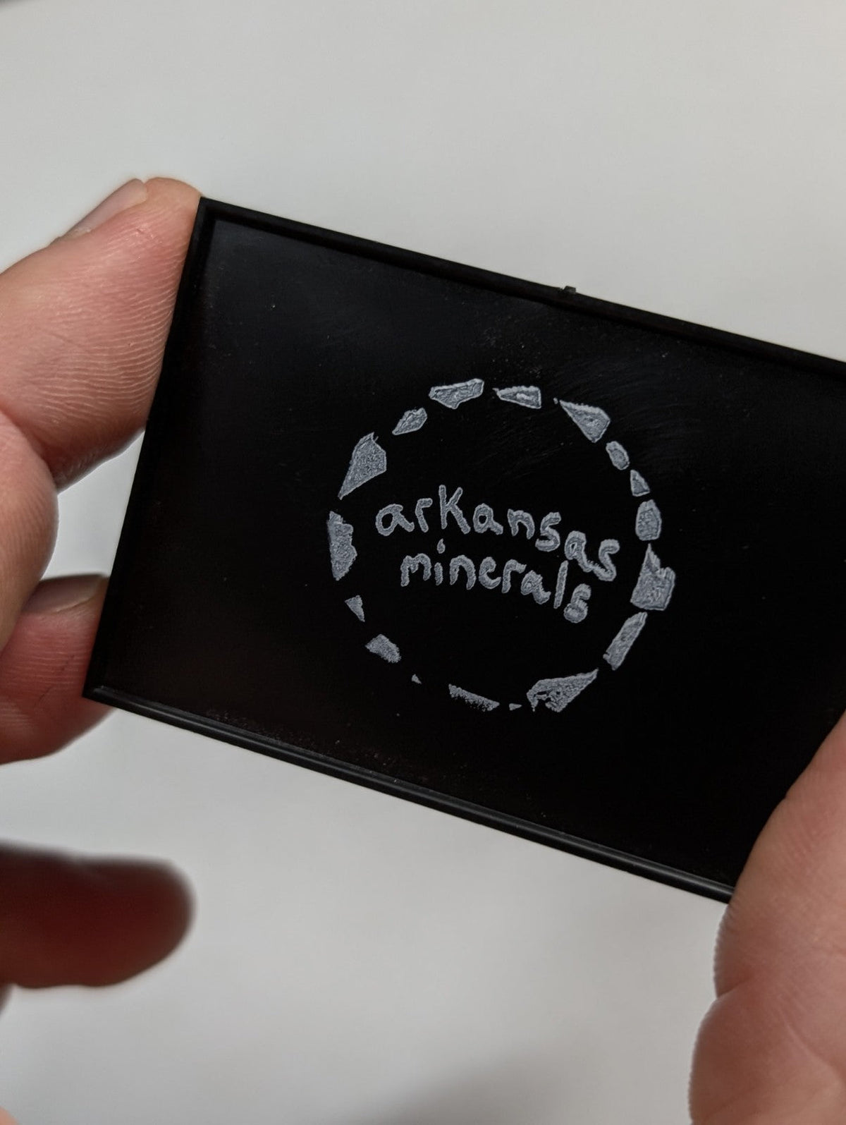 Black rectangular compact with silver engraved ’arkansas minerals’ logo surrounded by a circular pattern of stone shapes.