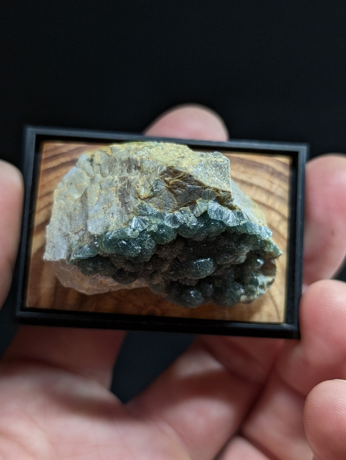 A framed mineral specimen featuring a greenish-blue crystalline formation with translucent, faceted crystals growing from a rougher, earthy matrix.