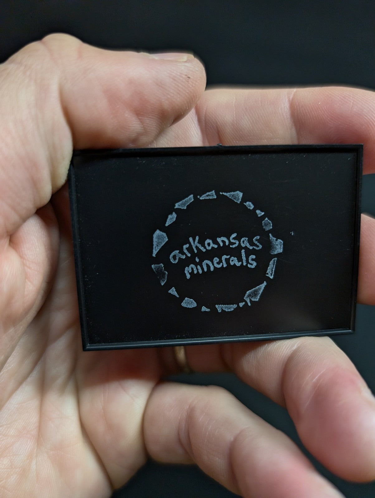 Black rectangular mineral sample case with ’arkansas minerals’ engraved in silver lettering surrounded by a circle of diamond-shaped motifs.