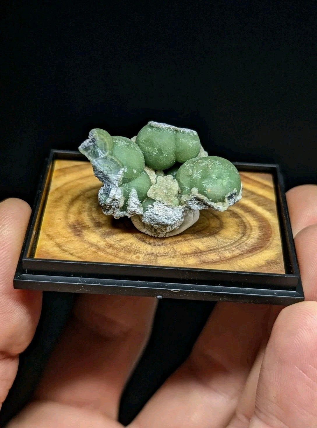 A polished green spherical mineral specimen with a crystalline, frosty texture rests on a wooden display base.