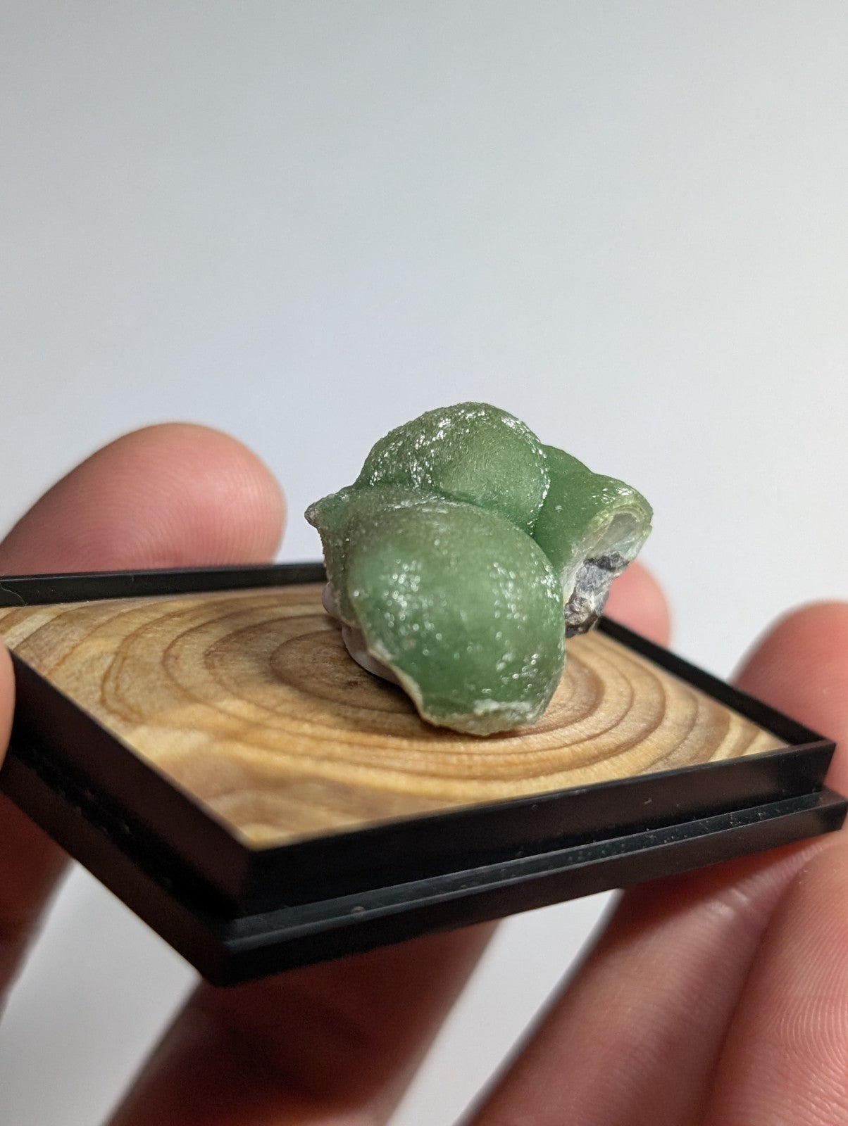 A translucent green mineral specimen with a smooth, rounded surface and subtle white speckles rests on a wooden display base.