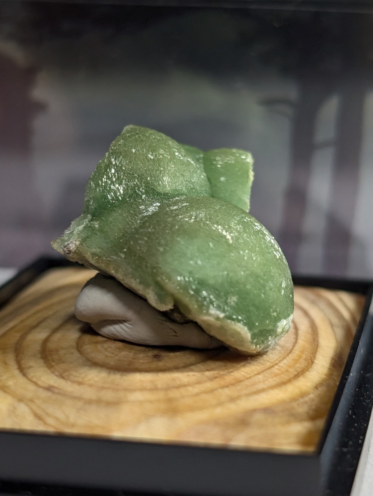 A smooth, translucent green stone with a glossy finish rests atop a smaller gray rock on a wooden display base.