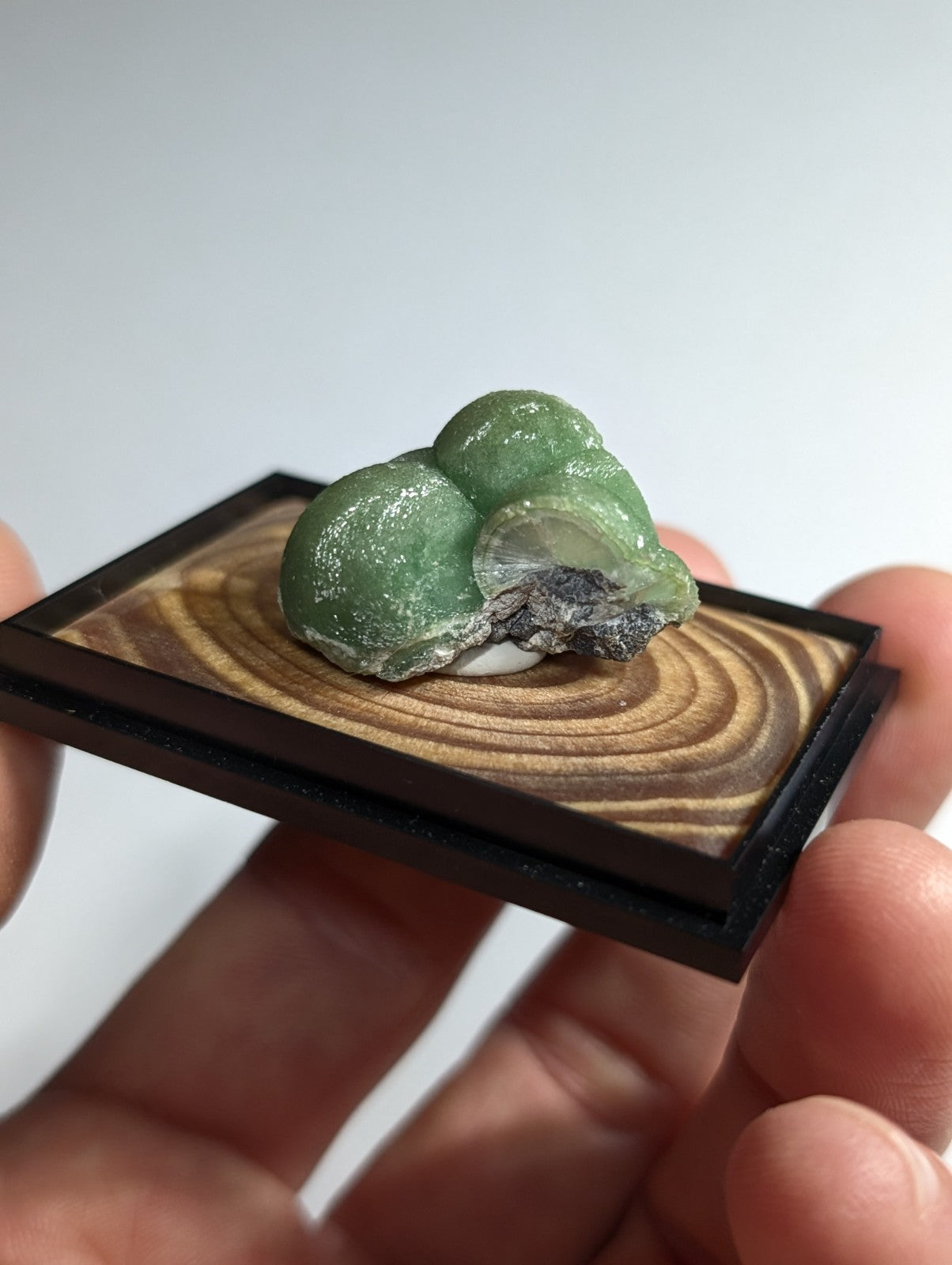 A polished green gemstone with a translucent, slightly frosted surface and natural mineral inclusions rests on a miniature wooden display tray.