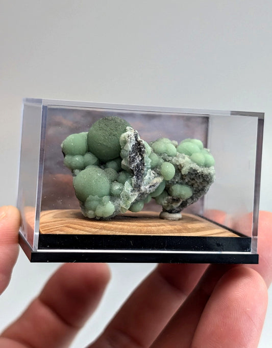 A green, bubbly mineral specimen with a mix of smooth spheres and rougher textures is displayed in a clear acrylic case on a wooden base.