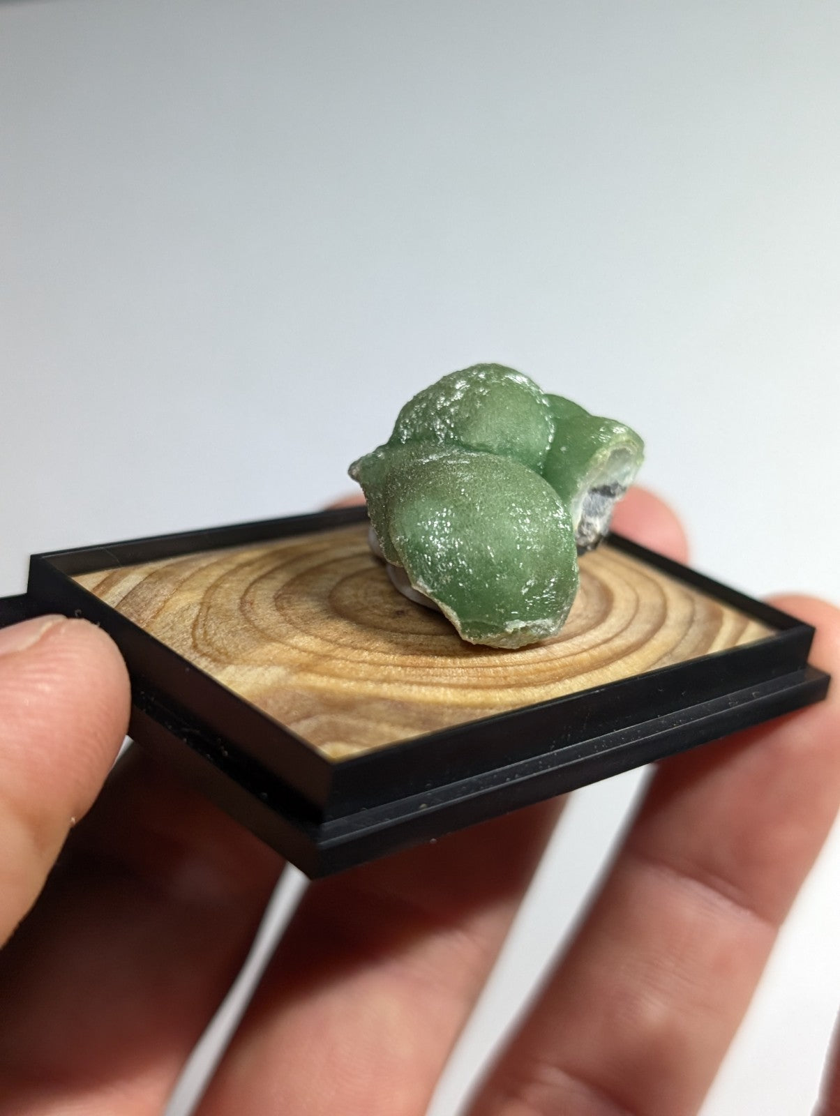 A smooth, translucent green mineral specimen with a slightly rough texture and natural irregular shape rests on a wooden display base.