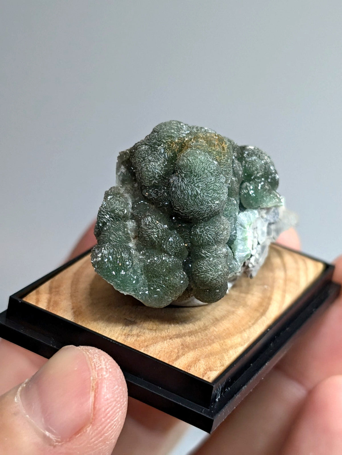 A green, bumpy mineral specimen with a speckled surface and hints of yellowish tones is displayed on a wooden base framed in black.